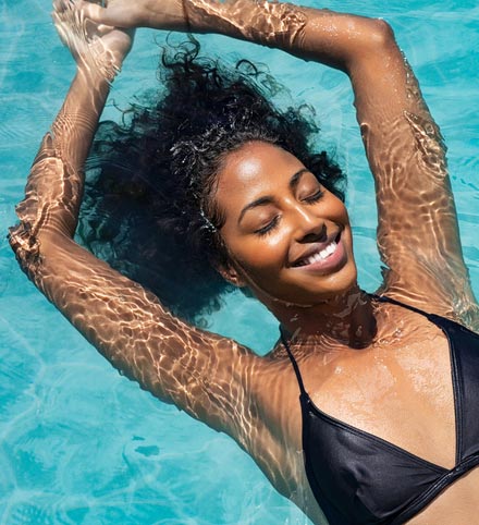 Woman with curly hair smiling and floating in a swimming pool with arms raised overhead